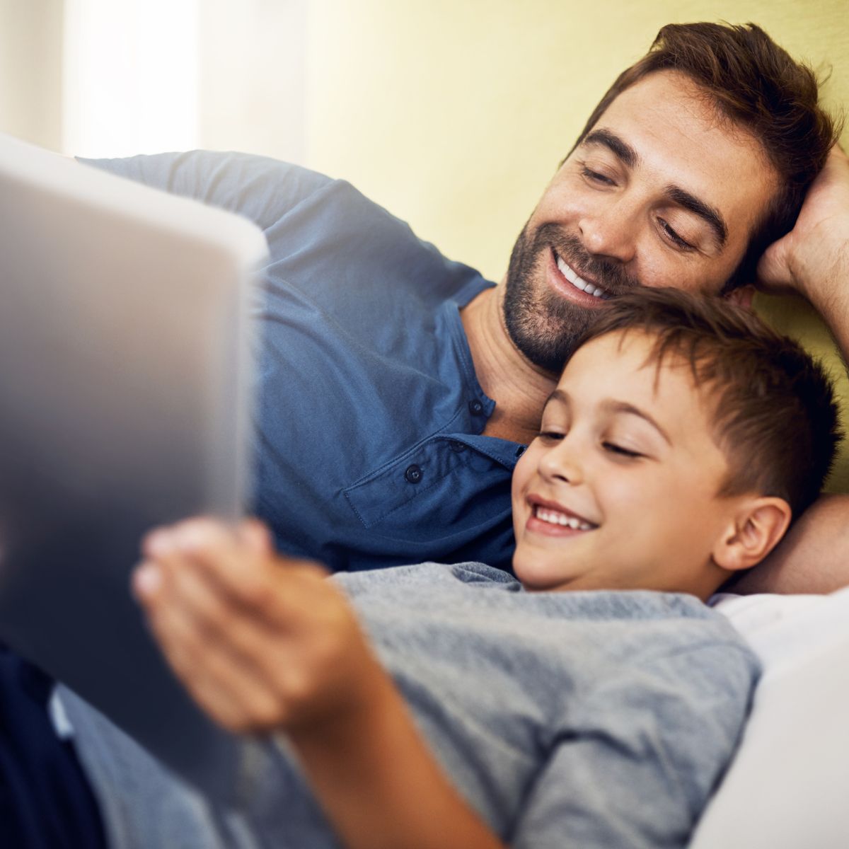 Digitale Medienkompetenz. Ein Mann in blauem Langarmhemd und ein Junge in grauem Pullover schauen gemeinsam auf ein Tablet. 