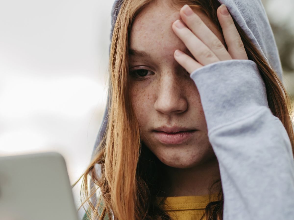 Jugendliche, die Opfer von Cybermobbing geworden ist, trägt einen grauen Kapuzenpullover und greift sich an die Stirn.