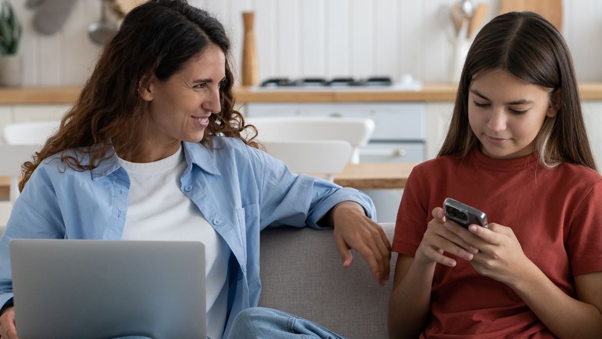 Eine Frau mit blauem Hemd und weißem T-Shirt und eine Jugendliche mit rotem T-Shirt sitzen auf einem Sofa. Im Hintergrund ist eine weiße Küchenzeile zu sehen. Die Jugendliche ist in ihr Smartphone vertieft, die Frau hat einen aufgeklappten Laptop auf dem Schoß. Die Jugendliche schaut auf ihr Handy, die Frau hat ihren Blick lächelnd auf die Jugendliche gerichtet.