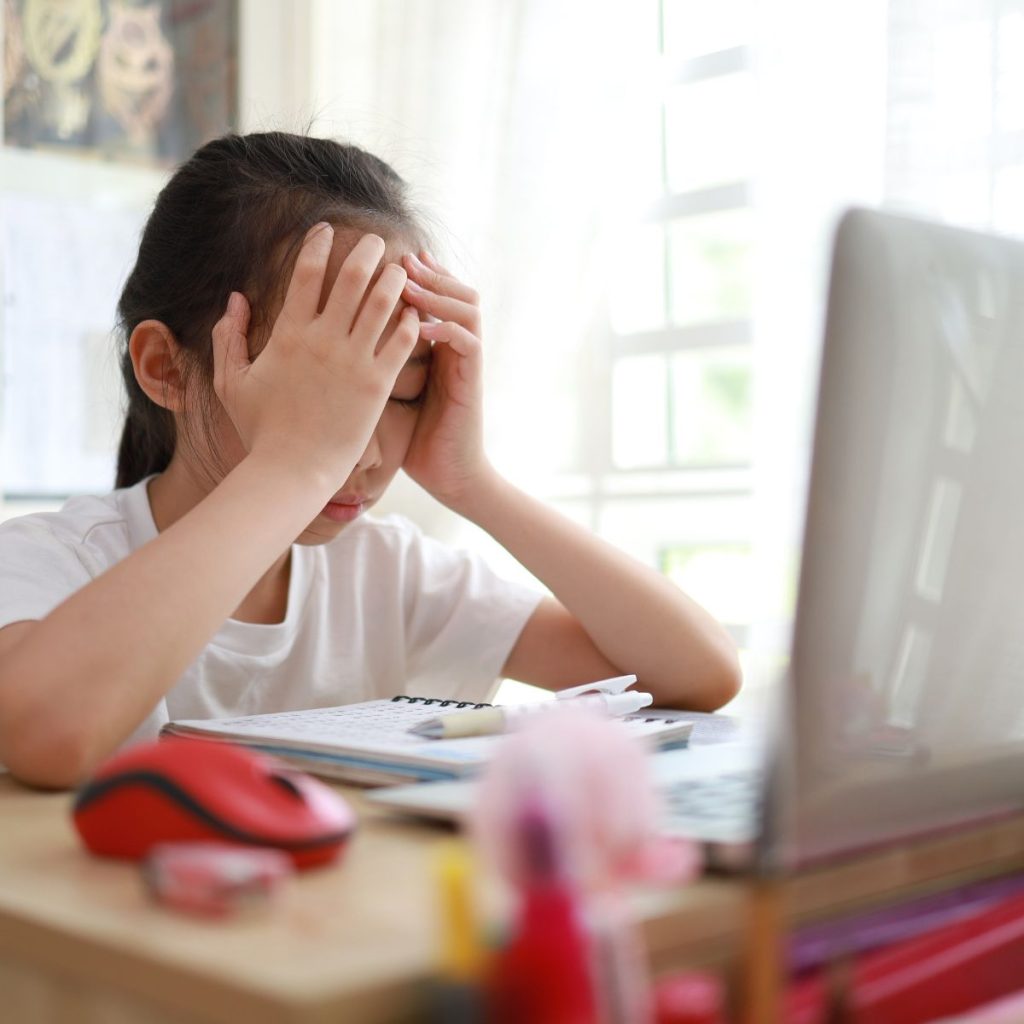 Ein Mädchen mit weißem T-Shirt schlägt die Hände über dem Kopf zusammen. Sie sitzt vor einem Laptop. Die dargestellte Situation zeigt ein Mädchen, das Opfer von Cyber-Mobbing geworden ist. 