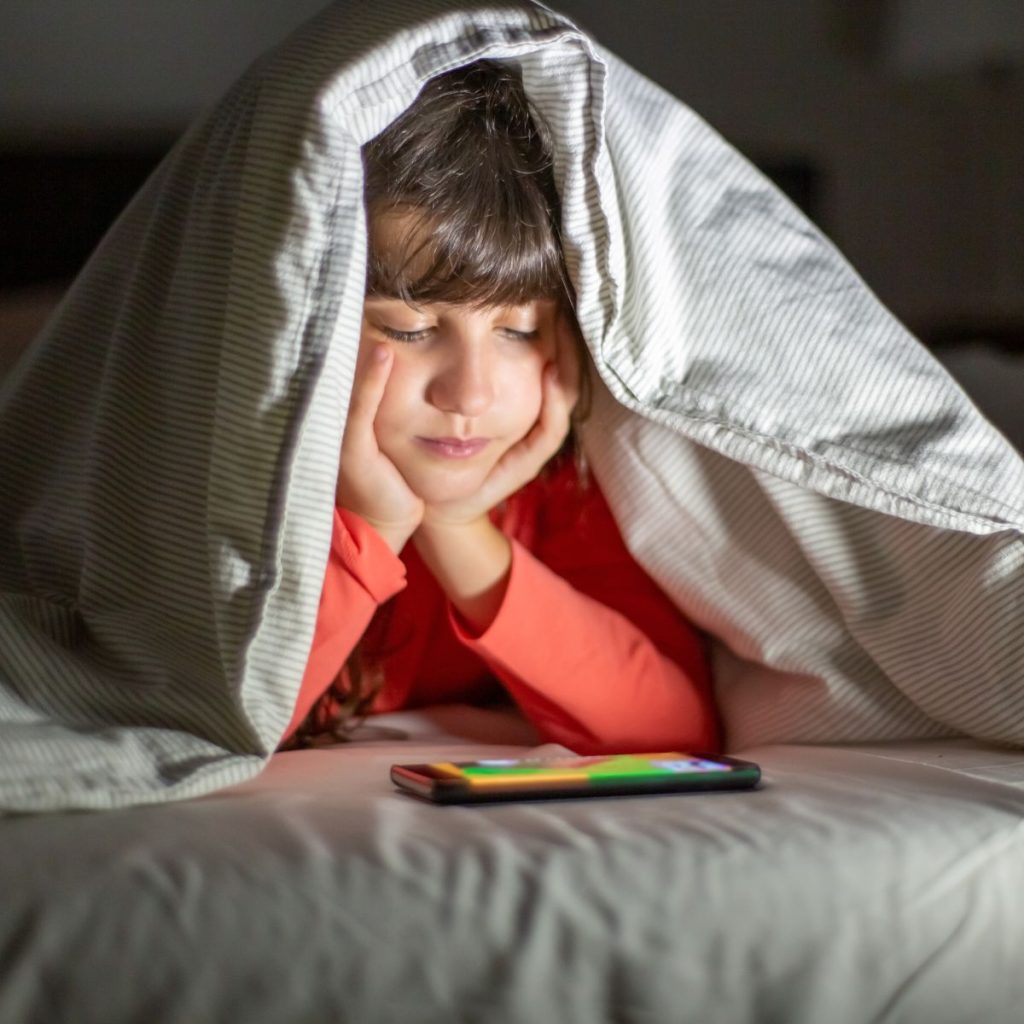 Ein Junge mit braunen Haaren und rotem Pullover liegt auf dem Bett, hat die Bettdecke über den Kopf gezogen und schaut auf ein Handy, das vor ihm auf dem Bett liegt.  