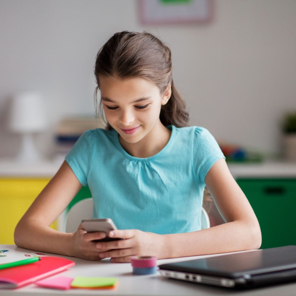 Ein Mädchen mit braunen Haaren und einem türkisfarbenen T-Shirt sitzt an einem Schreibtisch, vor ihr liegen Schulaufgaben und ein zugeklappter Laptop. Ihr Blick ist auf das Smartphone in ihren Händen gerichtet. 