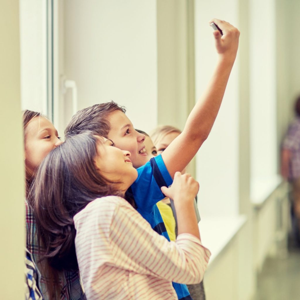 Fünf Kinder stehen am Fenster. Vier Mädchen und ein Junge. Der Junge in der Bildmitte hält ein Smartphone hoch und schießt ein Selfie von der ganzen Gruppe. Alle Kinder lächeln in die Kamera. 