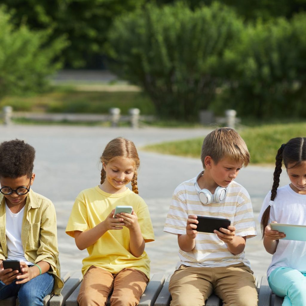 Auf einer Parkbank sitzen vier Kinder. Die Kinder sind im Grundschulalter. Es sind zwei Jungen und zwei Mädchen zu sehen. Alle Kinder haben ein Smartphone in der Hand. 